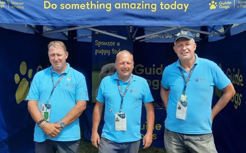 Three fundraisers stand in a row outside of a Guide Dogs stand