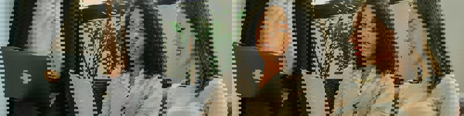 A group of three women are sitting on a sofa and looking at documents, and holding laptops in a professional setting. 