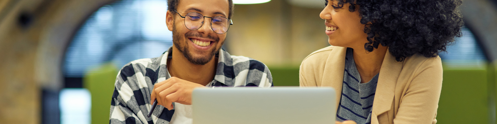 Two colleagues smiling and collaborating on a laptop in a bright, contemporary workspace with circular lighting fixtures.