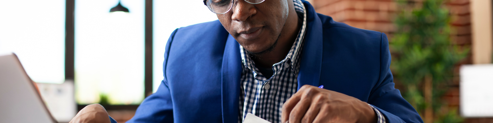 Person of colour wearing glasses and a blue suit jacket, concentrating whilst reviewing documents and working on a laptop in a bright office with exposed brick walls