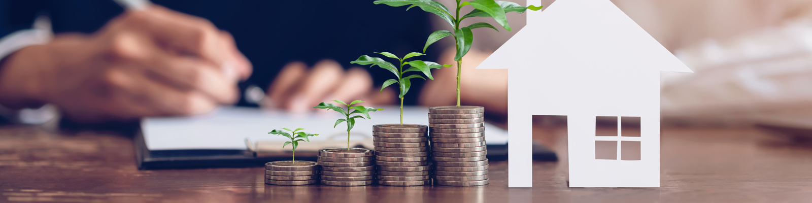 Stack of coins, each one higher than next with plant growing on top, and model house placed on the table.