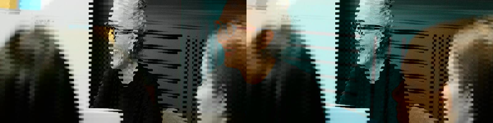 Older woman sat working on a laptop, leading a meeting. Two other professionals sit in the foreground out of focus. 