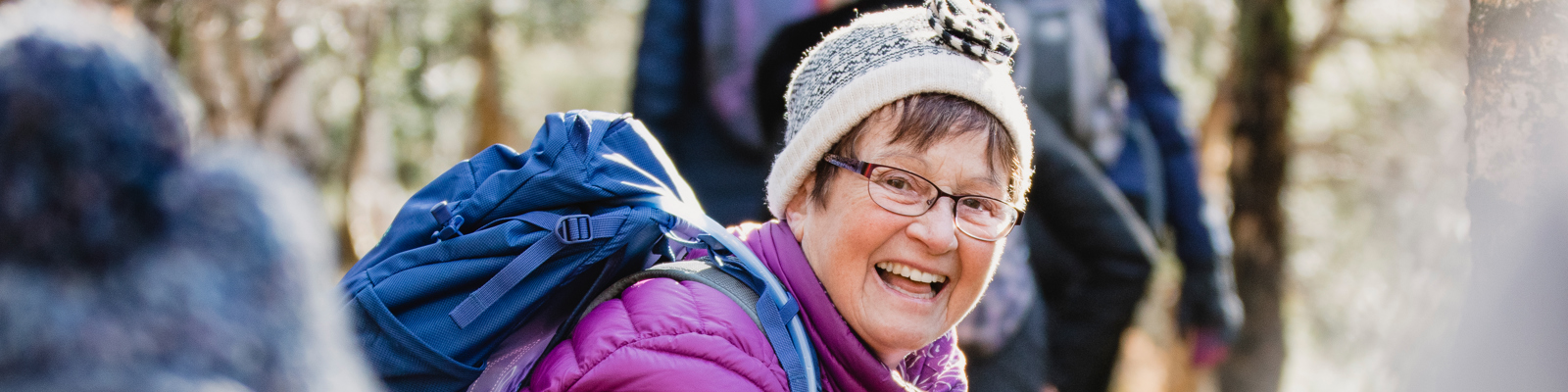 Photo of an older woman wearing a hat and hiking gear smiling looking back at the camera while during an event.