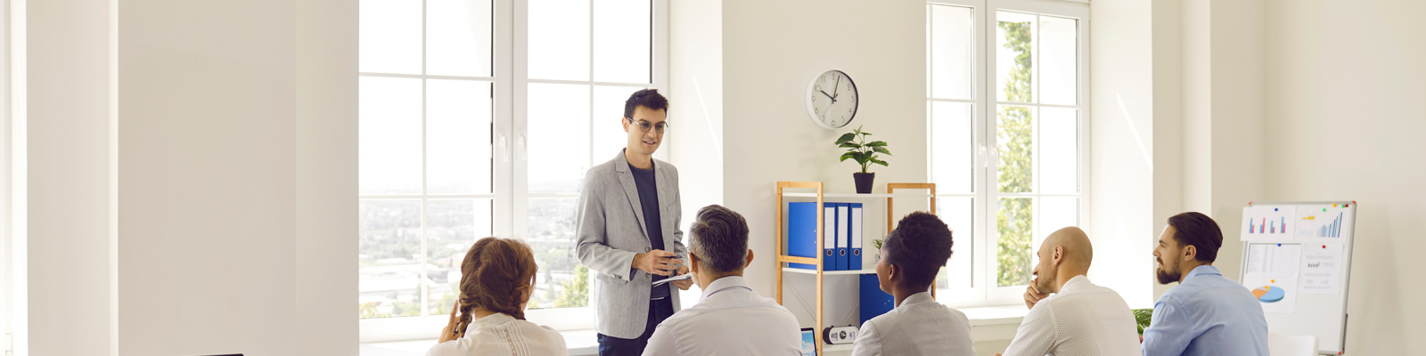 Young man in office speaks in front of business men and women sitting in row at table.