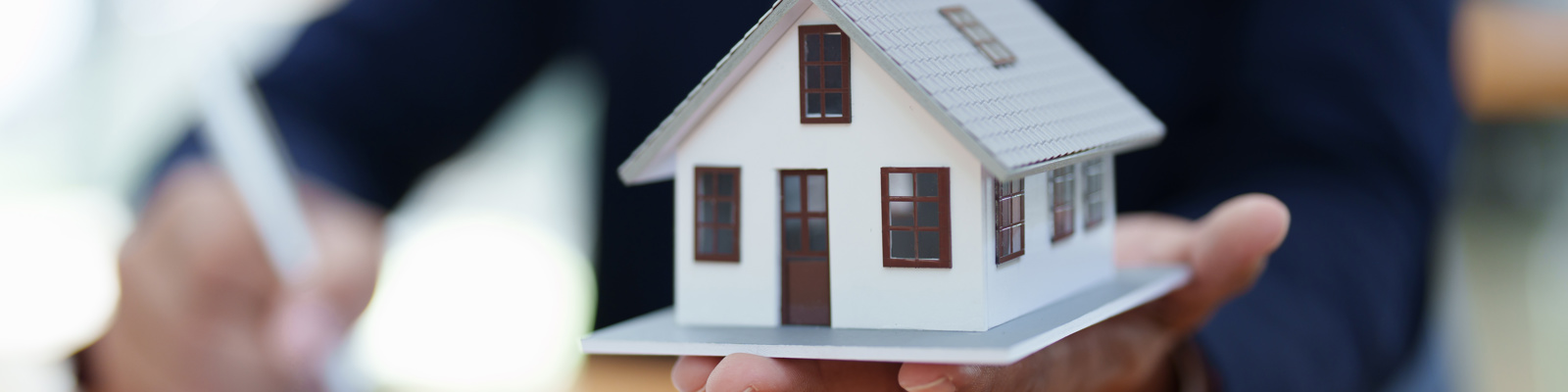 Photo of a person signing a document in the background while the foreground has the other hand holding out a minature size version of a house