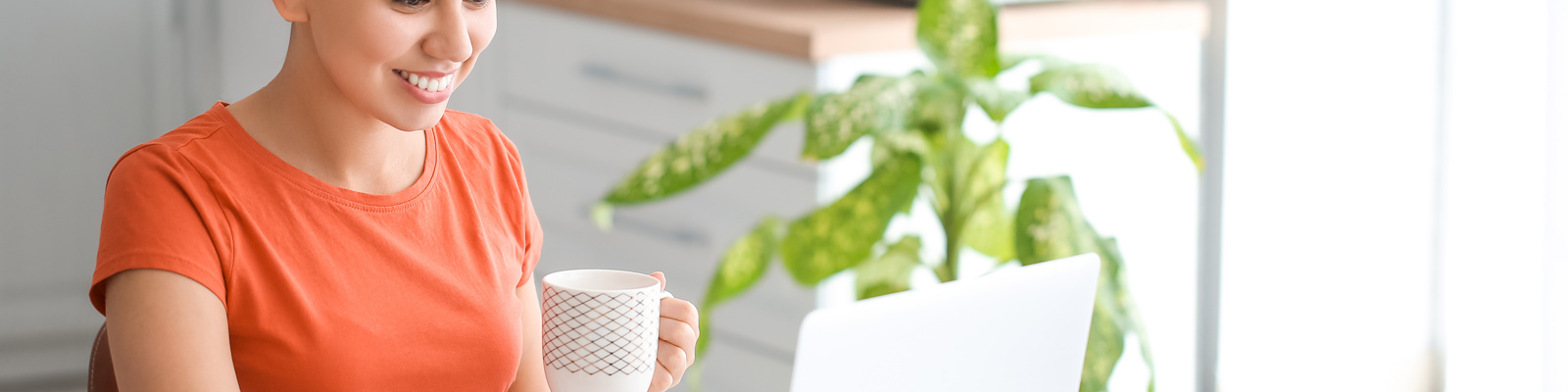 Woman with hair in a bun wearing an orange t-shirt, smiling whilst working on a laptop in a modern kitchen with plants nearby, holding a mug