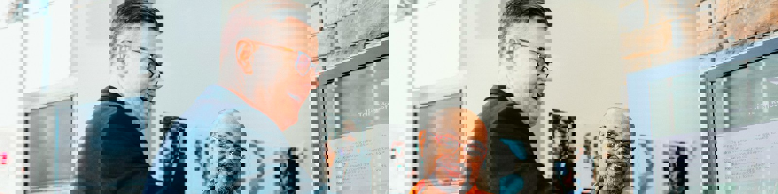 Two middle aged men stand at a desk, looking at something on a laptop.