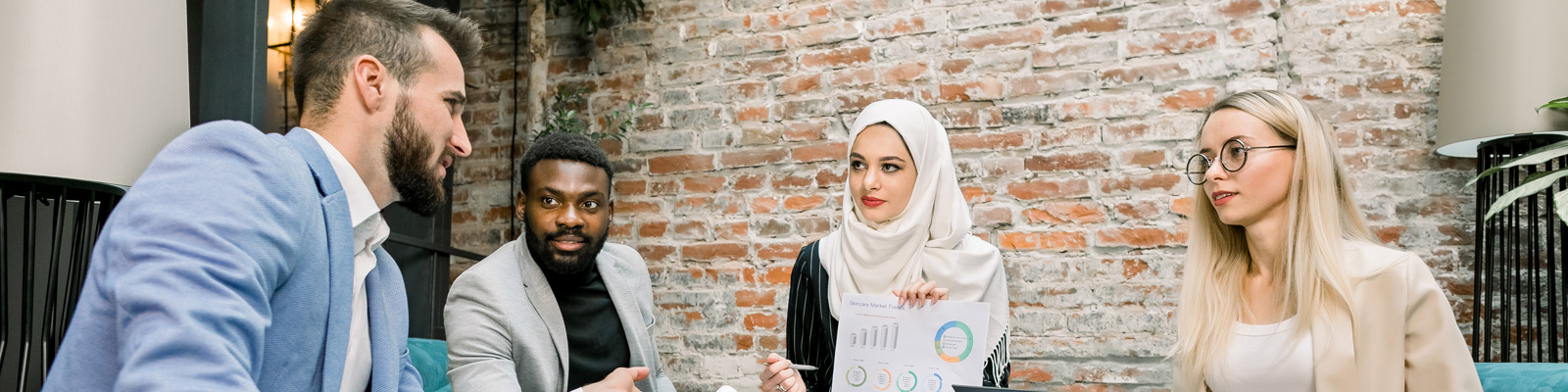 Diverse team of four professionals meeting in a modern office with exposed brick walls, seated on teal sofas, reviewing documents and data charts with laptops