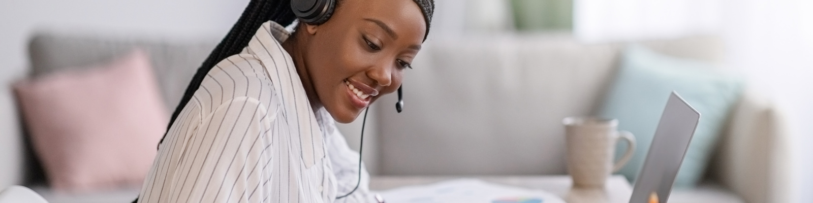 Black woman with braided hair in a striped shirt wearing headphones and smiling whilst writing notes and working from home