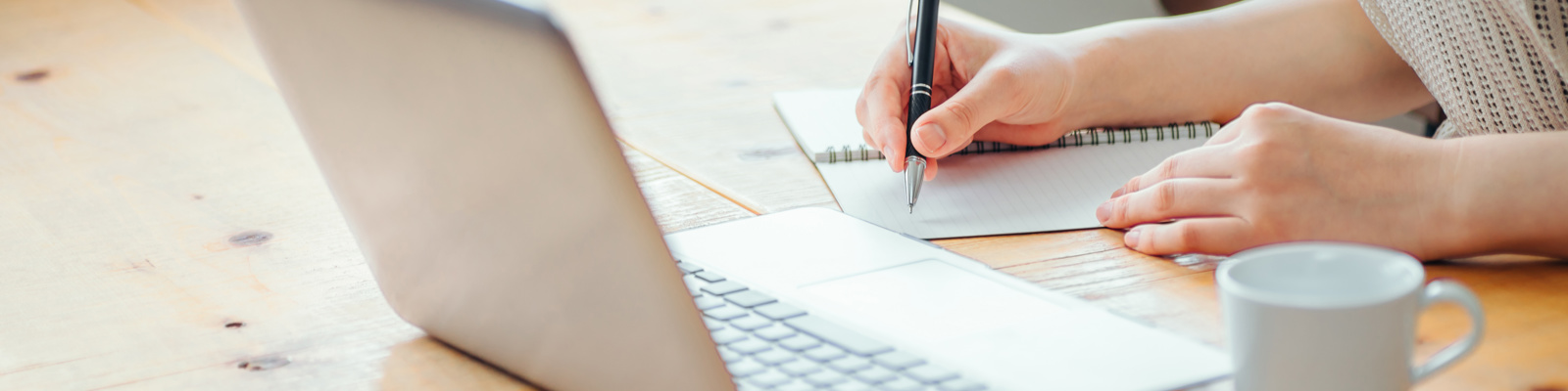 Photo of a woman's hand writing on a notebook suggesting writing a letter whilst looking at a laptop.