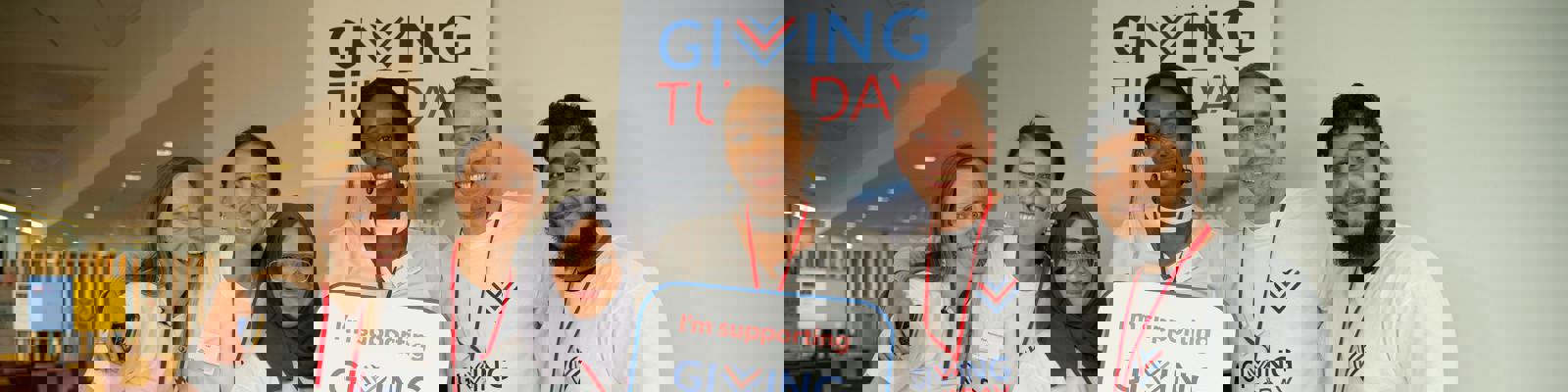 A Group of People Wearing Giving Tuesdat T-Shirts. One in the Middle is Holding a Sign Up That Says 'I'm Supporting Giving Tuesday'. The Giving Tuesday Logo is in the Background on the Wall.