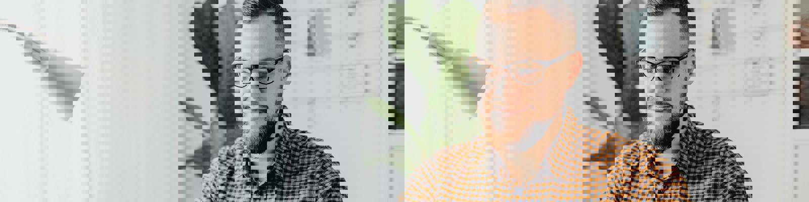 White man in a brown checked shirt, sitting in an office using a white laptop with a coffee mug by his side