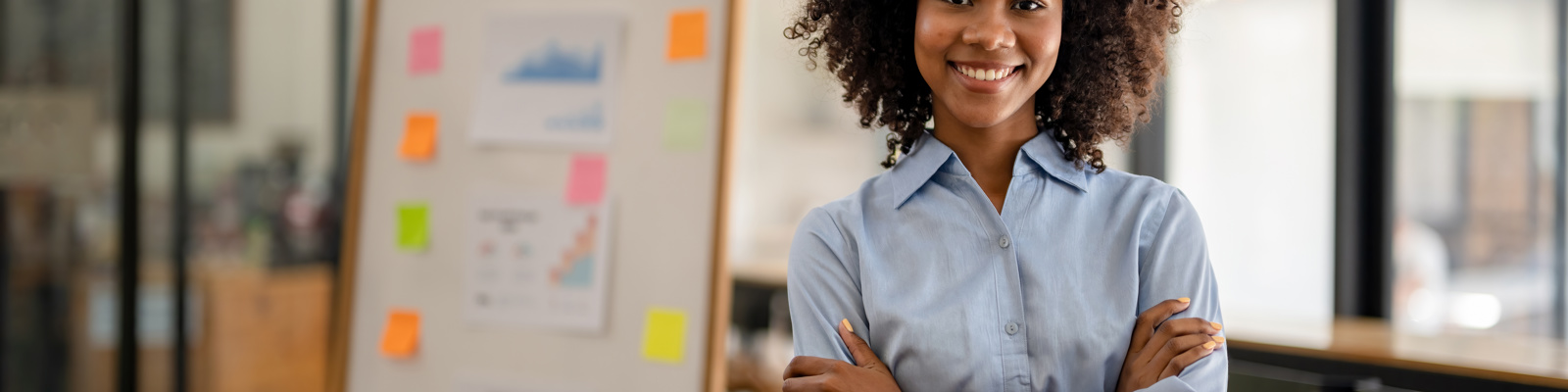 Confident woman in a light blue shirt standing with arms crossed beside a whiteboard with colorful project notes.