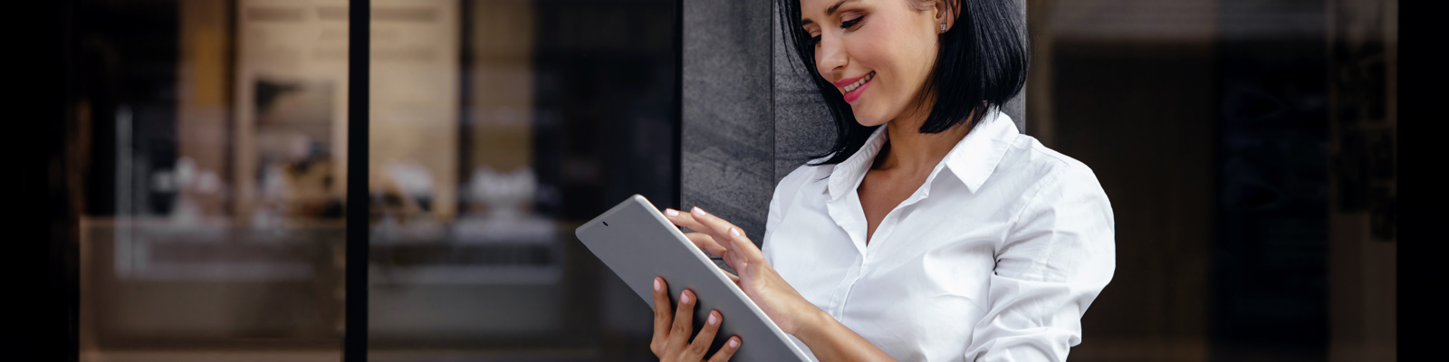 A woman standing outside, leaning against the dark pillar of a building and using a tablet computer